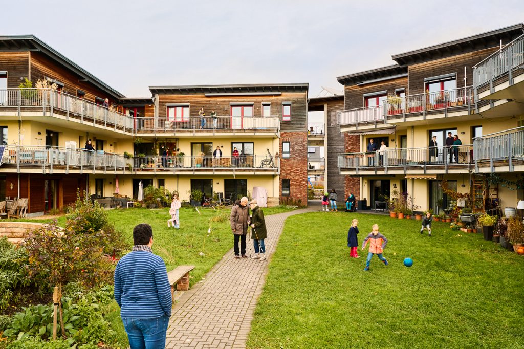 Innenhof des Mehrgenerationenwohnens: Menschen stehen auf Balkonen, laufen durch den Garten und Kinder spielen mit einem Ball